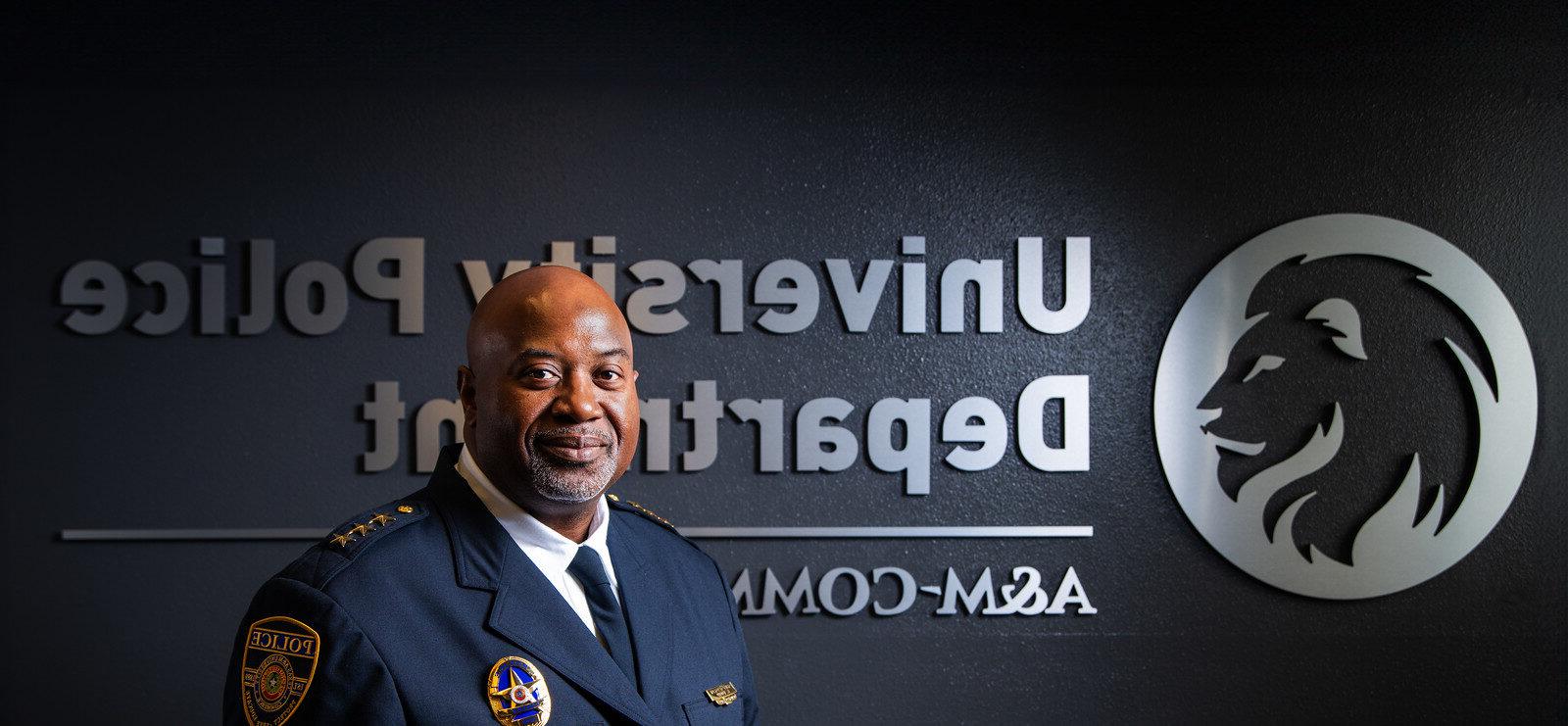 Chief Bryan Vaughn dressed in his dark blue, formal police uniform. He is standing in front of a dark wall with a silver embossed TAMUC lion head logo and the words University Police Department A&M-Commerce.